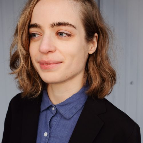 Veronique, a white non-binary person with side-swept, shoulder-length sandy blond hair, smiles while looking to the left. They are wearing a black blazer over a blue button-down shirt