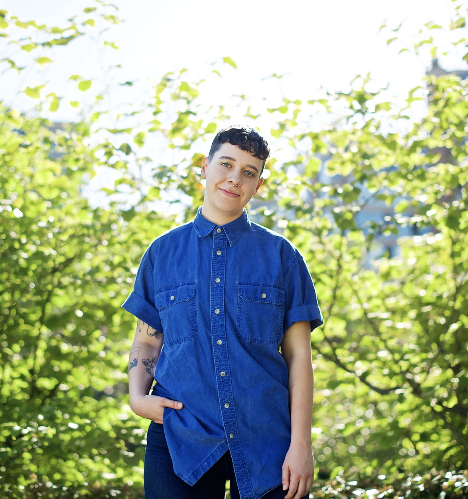Kit McKeown: wearing short-sleeve jean shirt, has brown hair an blue eyes and is similing at camera with one hand in pocket standing in front of green trees