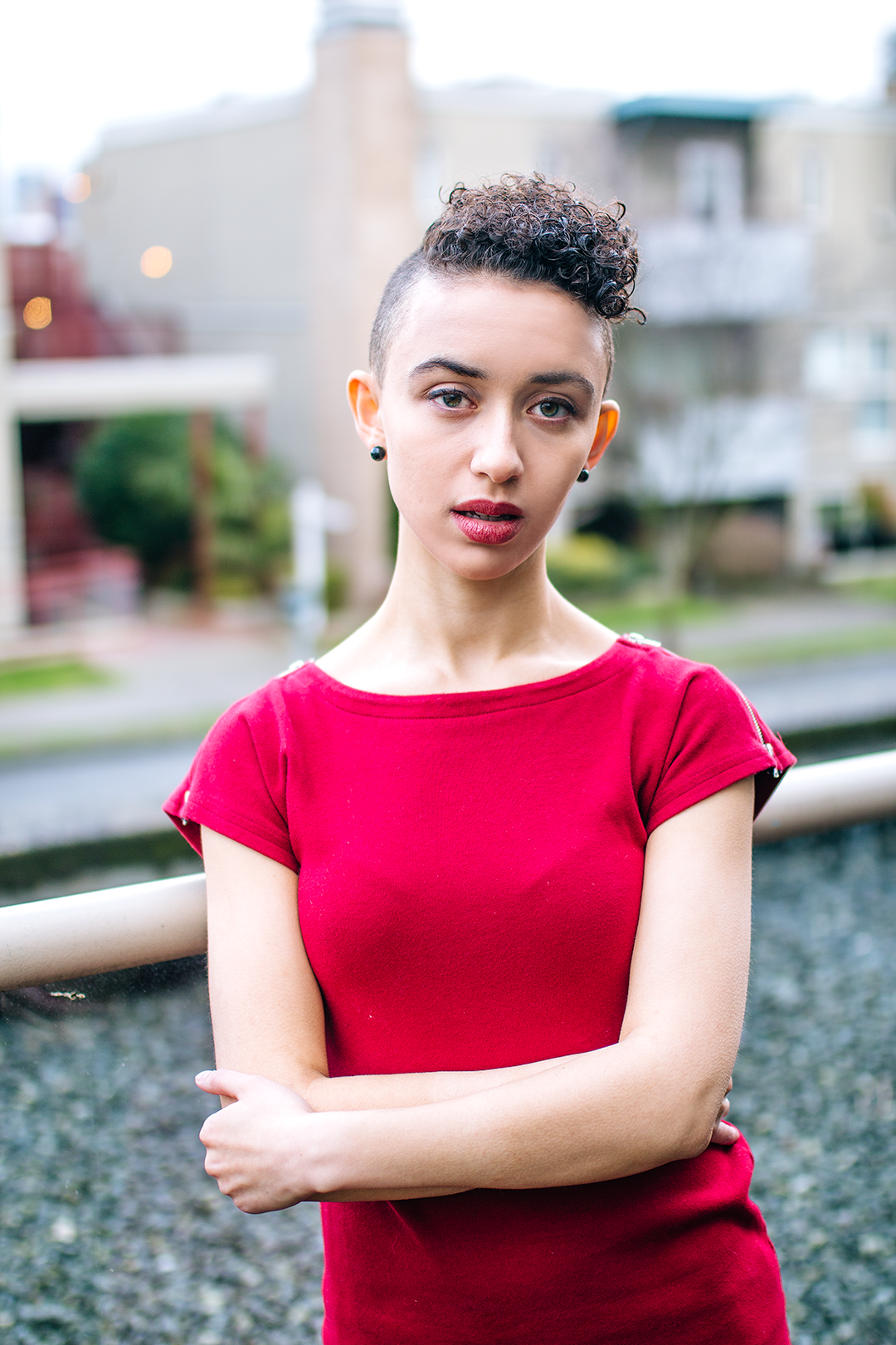 Lili Robinson, a genderfluid femme person in their mid-twenties, looks directly into the camera. They have light brown skin and short curly brown hair that is shaved on the sides, and they are wearing a red dress. In the background a gravel rooftop and apartment building across the road behind are blurred but visible.