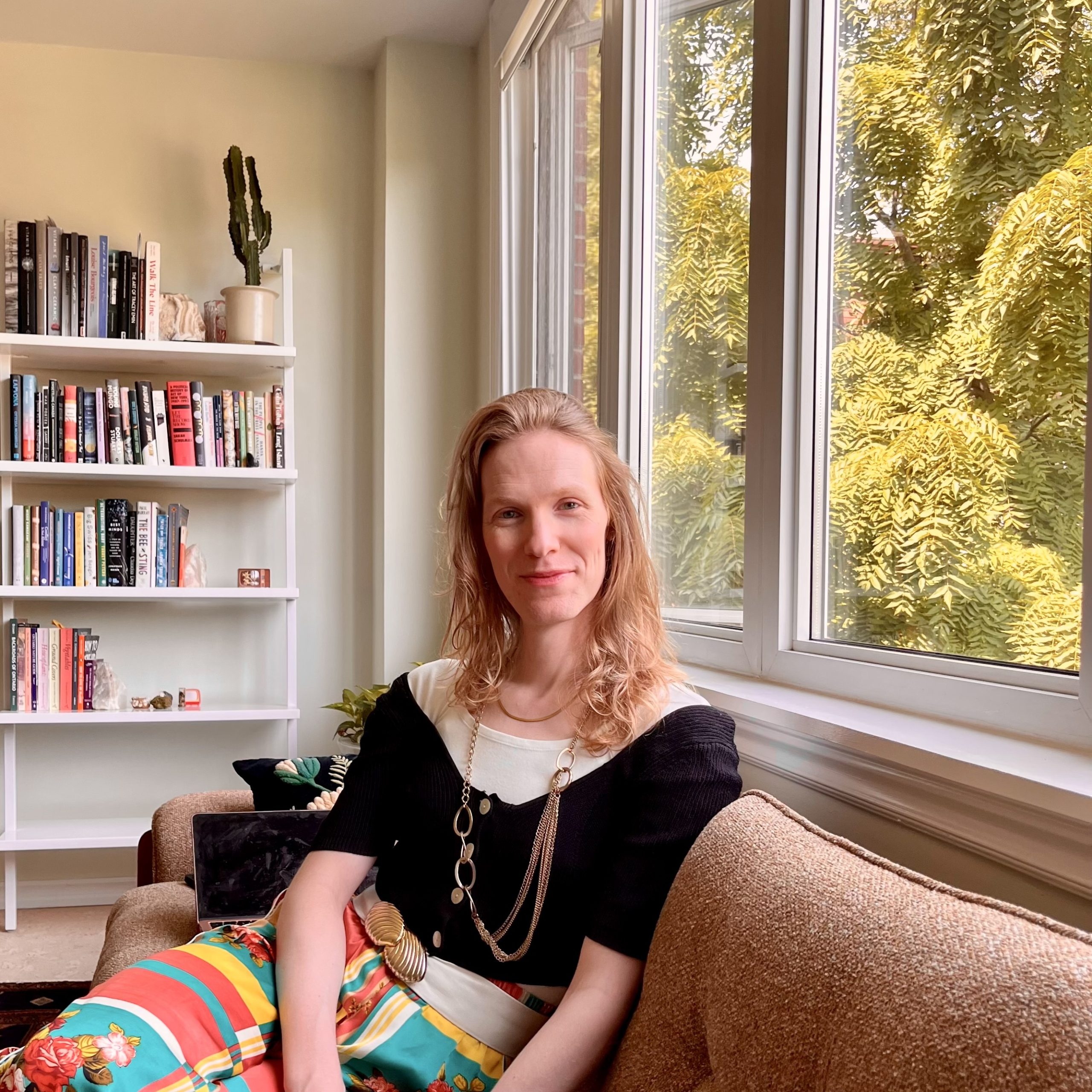Chrisina is seated on a taupe couch against a large, open window with trees on the outside on the right and a bookshelf with books to the left. She is smiling at the camera. She has shoulder length blonde hair and wearing a black, loose shirt over a white tshirt with a multi-coloured skirt accessorized with a long gold chain around her neck.