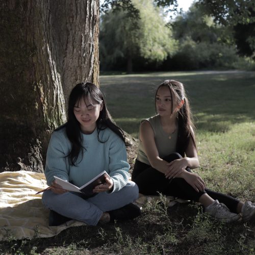 A photo of two young East Asian Canadian women sit at a base of a tree in a park of green lawns and trees. The central image for Before They Cut Down Our Tree by Karter Masuhara.