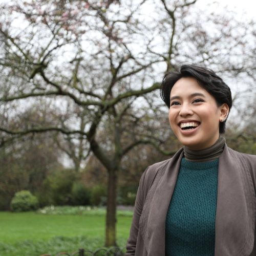 Renee is a Filipinx woman standing in a park with green grass and a tree in the background. She is smiling wide looking off to the left of the camera.