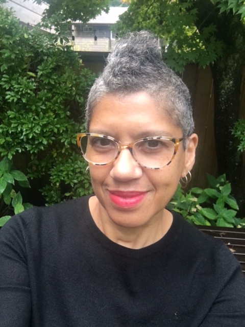 A black woman with short salt and pepper hair and styled in a mohawk smiles at the camera. She has tortoise glasses and red lipstick.